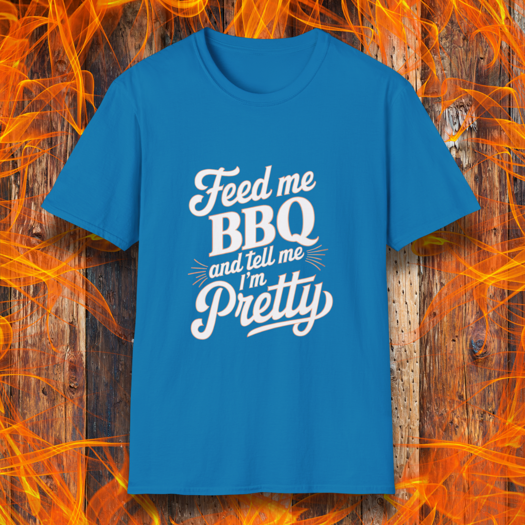 Sapphire T-shirt featuring a playful and bold typographic design that reads, "Feed me BBQ and tell me I'm Pretty." The text is styled in a mix of script and block fonts, with decorative accents emphasizing "BBQ" and "Pretty." The shirt is set against a rustic wooden background with vibrant orange flames, reinforcing the fiery BBQ theme.