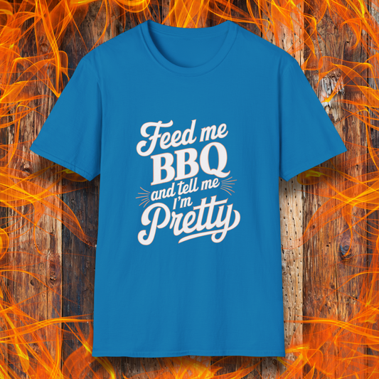 Sapphire T-shirt featuring a playful and bold typographic design that reads, "Feed me BBQ and tell me I'm Pretty." The text is styled in a mix of script and block fonts, with decorative accents emphasizing "BBQ" and "Pretty." The shirt is set against a rustic wooden background with vibrant orange flames, reinforcing the fiery BBQ theme.
