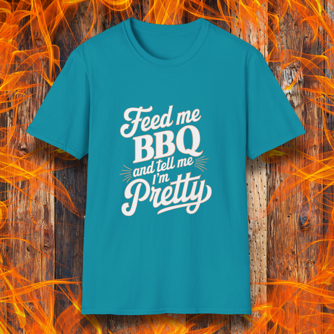 A Tropical Blue T-shirt featuring a playful and bold typographic design that reads, "Feed me BBQ and tell me I'm Pretty." The text is styled in a mix of script and block fonts, with decorative accents emphasizing "BBQ" and "Pretty." The shirt is set against a rustic wooden background with vibrant orange flames, reinforcing the fiery BBQ theme.