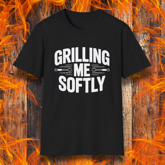 Black T-shirt with bold white text that says "Grilling Me Softly," featuring BBQ tool graphics between the words — set against a wood and flame background, perfect for grill lovers with a sense of humor.