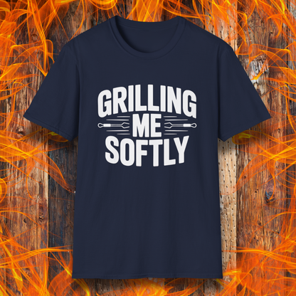 Navy T-shirt with bold white text that says "Grilling Me Softly," featuring BBQ tool graphics between the words — set against a wood and flame background, perfect for grill lovers with a sense of humor.