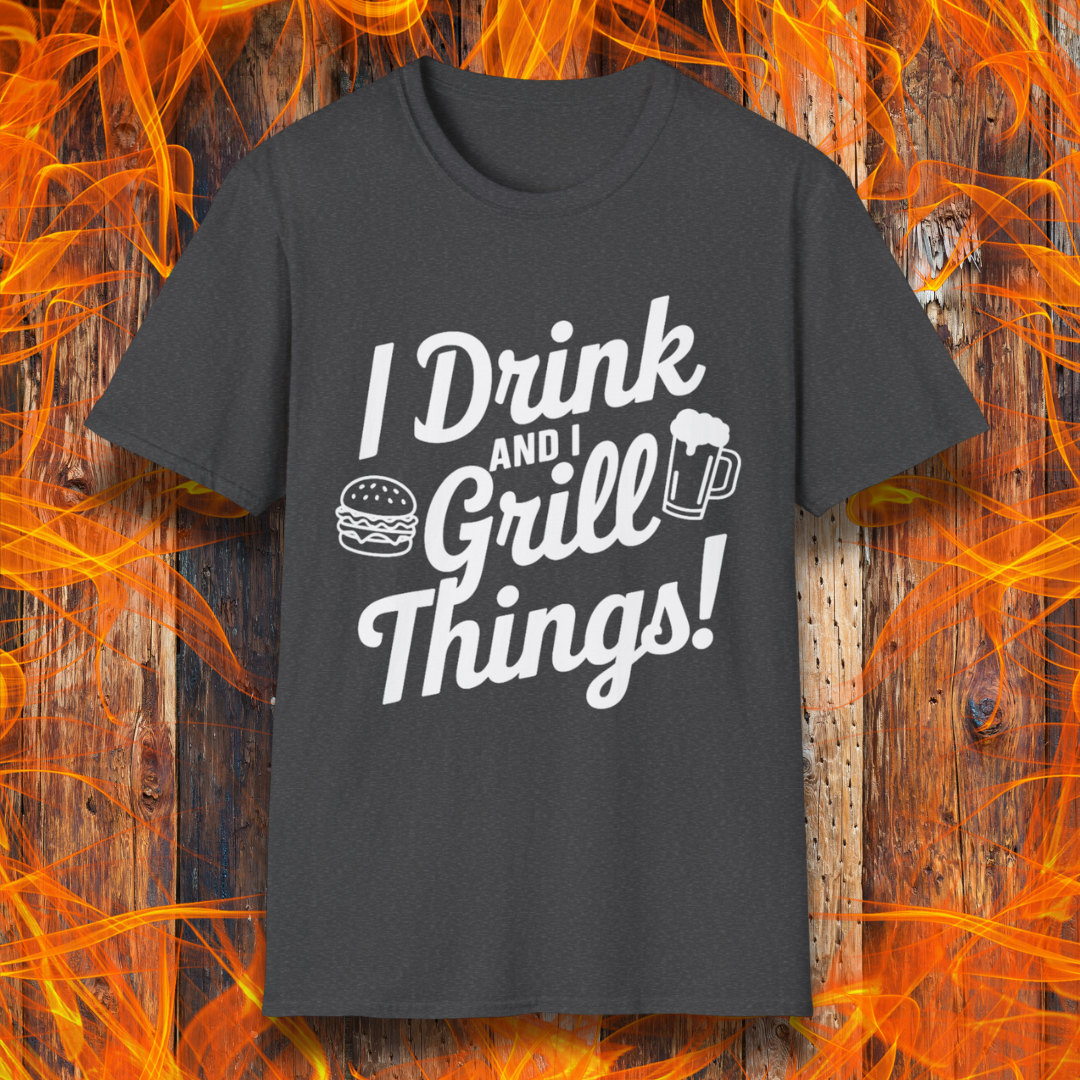 Dark Heather Grey T-shirt featuring a bold, black text design that reads, "I Drink and I Grill Things!" in a playful, script-style font. The design includes small illustrations of a burger and a frothy beer mug, emphasizing a fun and laid-back grilling theme. The background showcases a rustic wooden surface with bright orange flames, enhancing the fiery BBQ aesthetic.