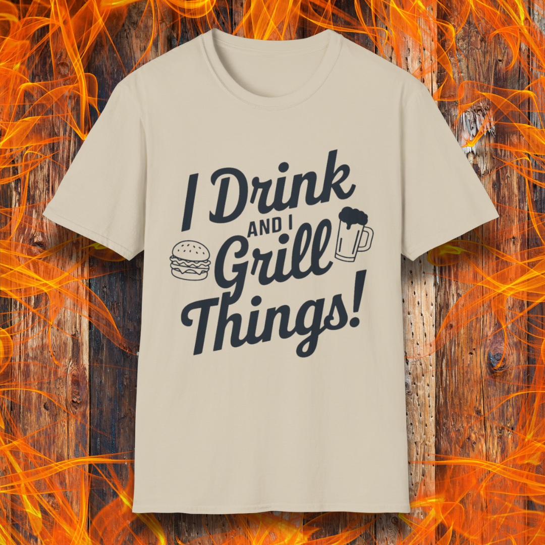 Sand or Tan Colored T-shirt featuring a bold, black text design that reads, "I Drink and I Grill Things!" in a playful, script-style font. The design includes small illustrations of a burger and a frothy beer mug, emphasizing a fun and laid-back grilling theme. The background showcases a rustic wooden surface with bright orange flames, enhancing the fiery BBQ aesthetic.
