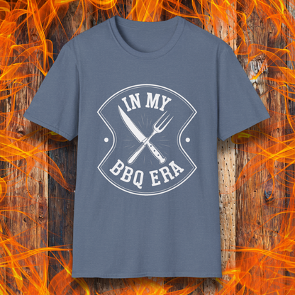 Heather Indigo T-shirt featuring a bold black design with the phrase "IN MY BBQ ERA" inside a shield-like emblem. At the center of the emblem, there is a crossed knife and grilling fork, symbolizing BBQ culture. The background showcases a rustic wooden texture with bright orange flames, adding a fiery and intense grilling aesthetic.