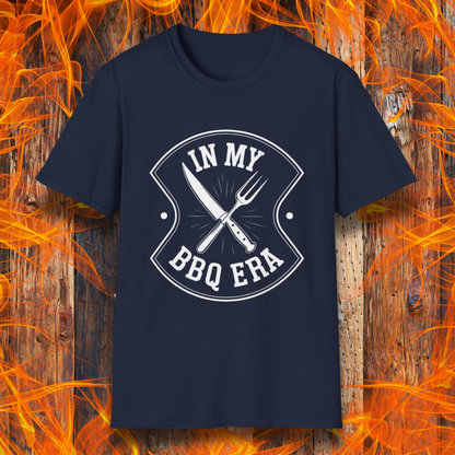 Navy T-shirt featuring a bold black design with the phrase "IN MY BBQ ERA" inside a shield-like emblem. At the center of the emblem, there is a crossed knife and grilling fork, symbolizing BBQ culture. The background showcases a rustic wooden texture with bright orange flames, adding a fiery and intense grilling aesthetic.