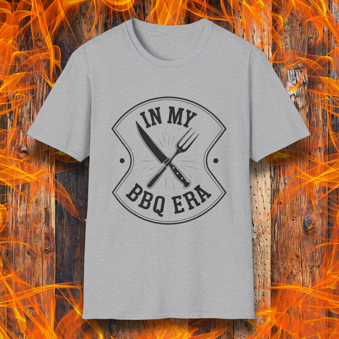 Grey T-shirt featuring a bold black design with the phrase "IN MY BBQ ERA" inside a shield-like emblem. At the center of the emblem, there is a crossed knife and grilling fork, symbolizing BBQ culture. The background showcases a rustic wooden texture with bright orange flames, adding a fiery and intense grilling aesthetic.