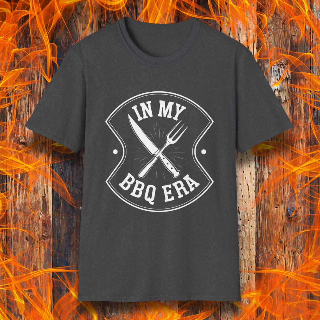 Dark Heather T-shirt featuring a bold black design with the phrase "IN MY BBQ ERA" inside a shield-like emblem. At the center of the emblem, there is a crossed knife and grilling fork, symbolizing BBQ culture. The background showcases a rustic wooden texture with bright orange flames, adding a fiery and intense grilling aesthetic.