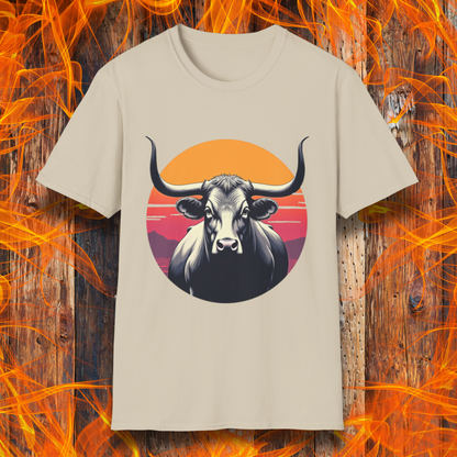 Sand T-shirt featuring a striking design of a longhorn bull staring forward with an intense gaze. The bull is set against a circular backdrop of a sunset with warm orange, red, and pink hues, along with silhouetted mountains in the background. The design conveys strength, confidence, and a bold Western aesthetic, appealing to BBQ and cattle enthusiasts.
