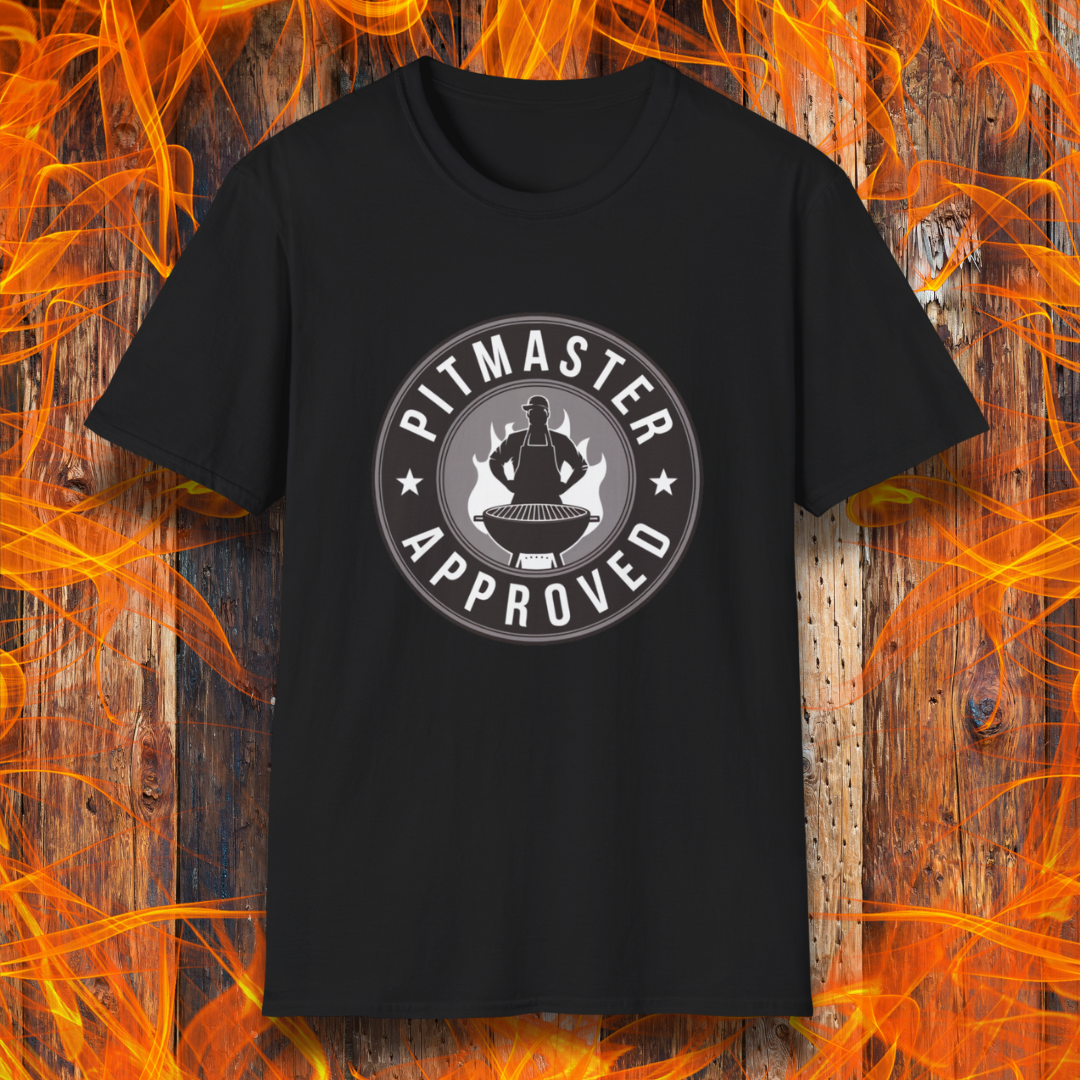 Black T-shirt featuring a circular badge-style design that reads "Pitmaster Approved." The center of the badge showcases a flaming grill with a silhouette of a sauce bottle above it, surrounded by stars and bold typography. The shirt is displayed against a rustic wooden background with bright orange flames, enhancing the BBQ theme.