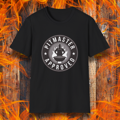 Black T-shirt featuring a circular badge-style design that reads "Pitmaster Approved." The center of the badge showcases a flaming grill with a silhouette of a sauce bottle above it, surrounded by stars and bold typography. The shirt is displayed against a rustic wooden background with bright orange flames, enhancing the BBQ theme.