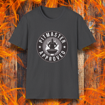 Dark heather T-shirt featuring a circular badge-style design that reads "Pitmaster Approved." The center of the badge showcases a flaming grill with a silhouette of a sauce bottle above it, surrounded by stars and bold typography. The shirt is displayed against a rustic wooden background with bright orange flames, enhancing the BBQ theme.