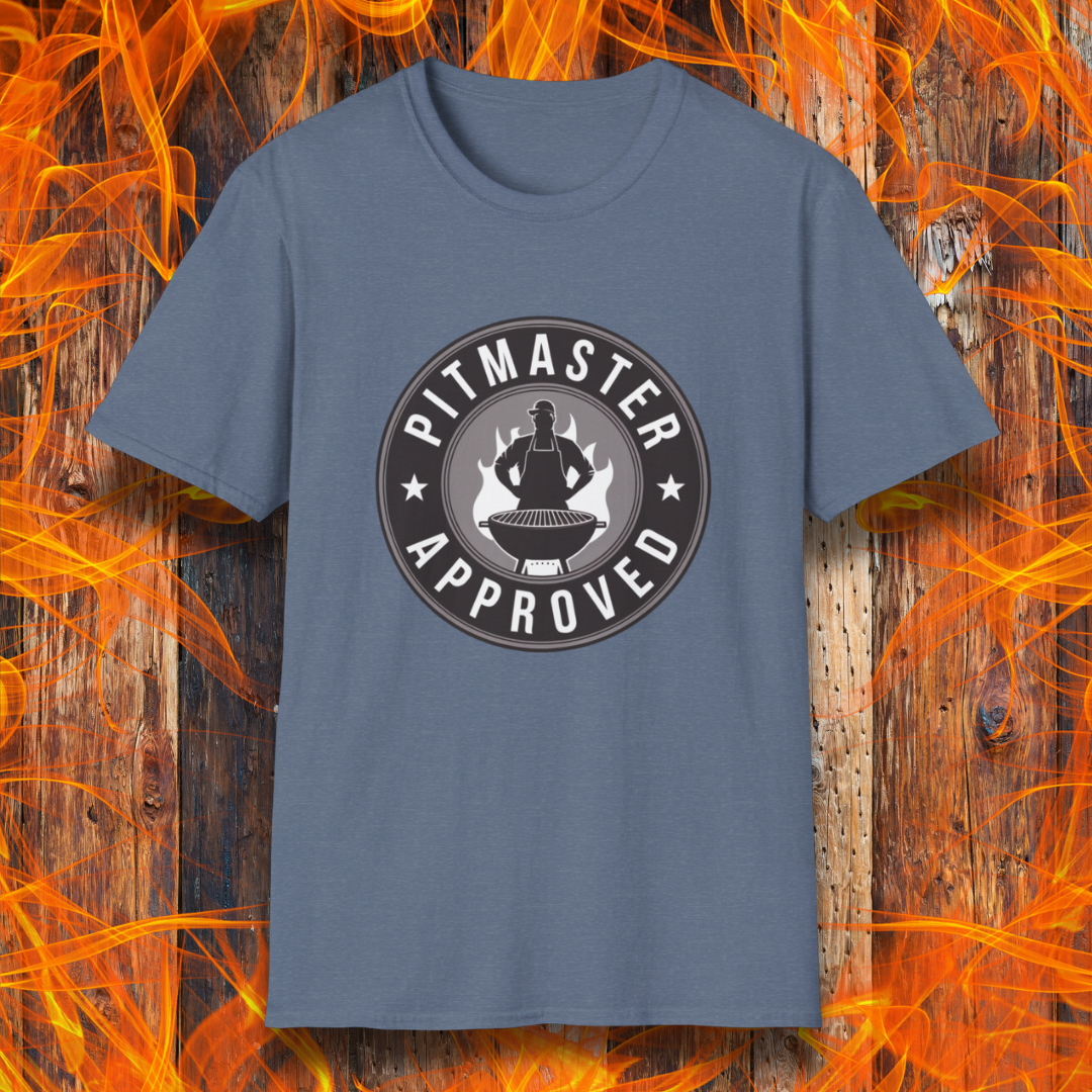 Heather Indigo  T-shirt featuring a circular badge-style design that reads "Pitmaster Approved." The center of the badge showcases a flaming grill with a silhouette of a sauce bottle above it, surrounded by stars and bold typography. The shirt is displayed against a rustic wooden background with bright orange flames, enhancing the BBQ theme.