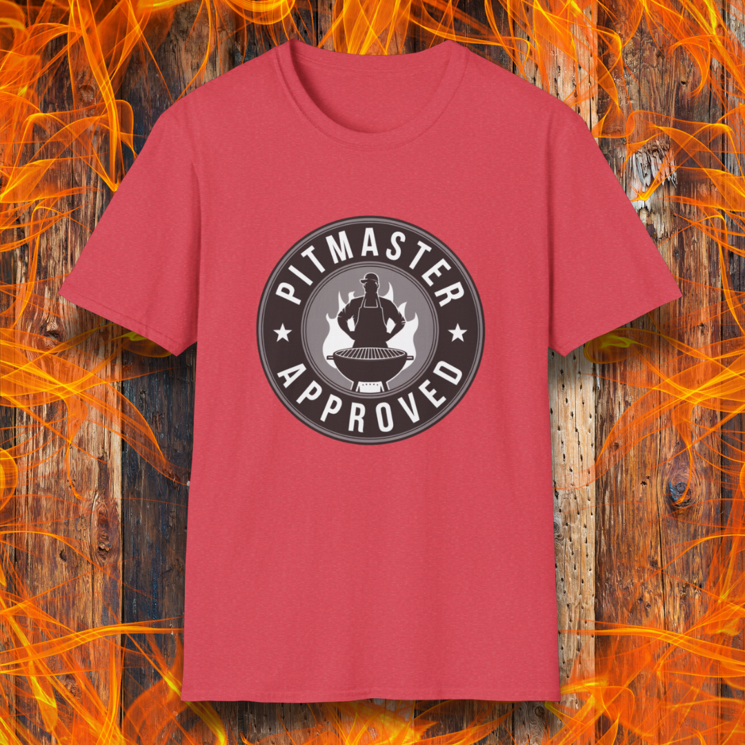 Heather red T-shirt featuring a circular badge-style design that reads "Pitmaster Approved." The center of the badge showcases a flaming grill with a silhouette of a sauce bottle above it, surrounded by stars and bold typography. The shirt is displayed against a rustic wooden background with bright orange flames, enhancing the BBQ theme.