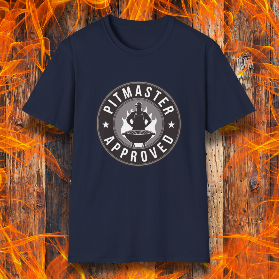 Navy T-shirt featuring a circular badge-style design that reads "Pitmaster Approved." The center of the badge showcases a flaming grill with a silhouette of a sauce bottle above it, surrounded by stars and bold typography. The shirt is displayed against a rustic wooden background with bright orange flames, enhancing the BBQ theme.