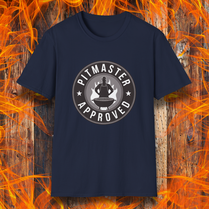 Navy T-shirt featuring a circular badge-style design that reads "Pitmaster Approved." The center of the badge showcases a flaming grill with a silhouette of a sauce bottle above it, surrounded by stars and bold typography. The shirt is displayed against a rustic wooden background with bright orange flames, enhancing the BBQ theme.