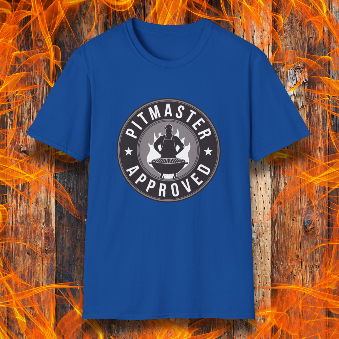 Royal Blue T-shirt featuring a circular badge-style design that reads "Pitmaster Approved." The center of the badge showcases a flaming grill with a silhouette of a sauce bottle above it, surrounded by stars and bold typography. The shirt is displayed against a rustic wooden background with bright orange flames, enhancing the BBQ theme.