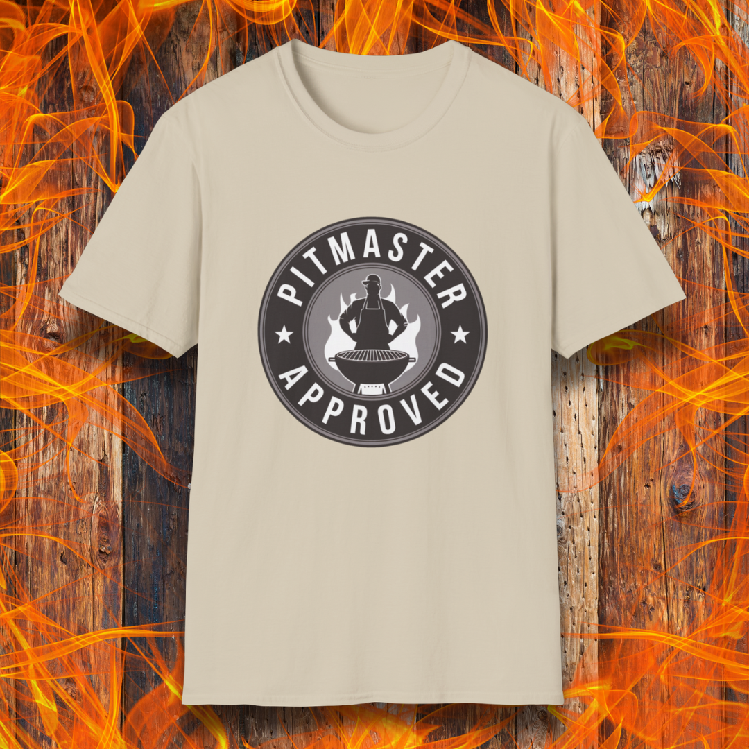 Sand T-shirt featuring a circular badge-style design that reads "Pitmaster Approved." The center of the badge showcases a flaming grill with a silhouette of a sauce bottle above it, surrounded by stars and bold typography. The shirt is displayed against a rustic wooden background with bright orange flames, enhancing the BBQ theme.