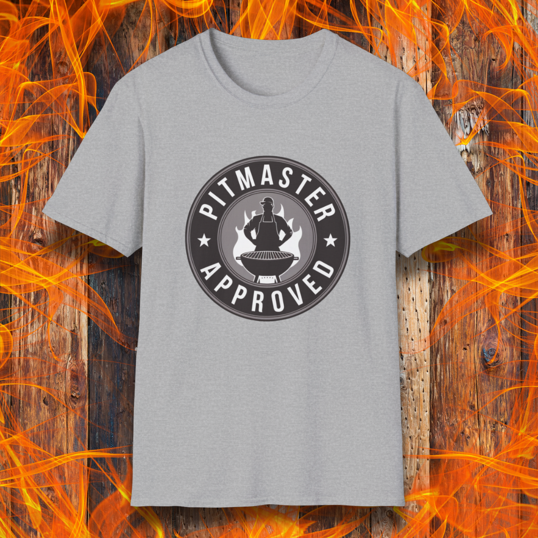 Sport grey T-shirt featuring a circular badge-style design that reads "Pitmaster Approved." The center of the badge showcases a flaming grill with a silhouette of a sauce bottle above it, surrounded by stars and bold typography. The shirt is displayed against a rustic wooden background with bright orange flames, enhancing the BBQ theme.