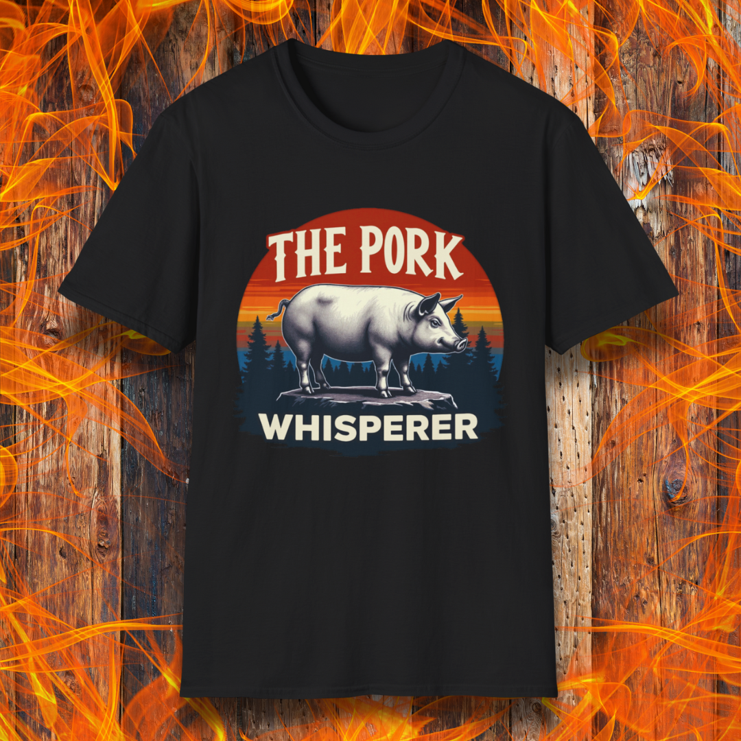 Black T-shirt featuring a bold and humorous BBQ-themed design. The centerpiece is a detailed pig standing on a wooden stump, framed by a circular emblem with a rustic sunset background in shades of orange, red, and yellow, along with pine tree silhouettes. Above the pig, the text reads "THE PORK WHISPERER" in bold, vintage-style lettering. This design is perfect for BBQ enthusiasts, pitmasters, and pork lovers.