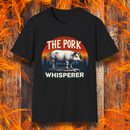 Black T-shirt featuring a bold and humorous BBQ-themed design. The centerpiece is a detailed pig standing on a wooden stump, framed by a circular emblem with a rustic sunset background in shades of orange, red, and yellow, along with pine tree silhouettes. Above the pig, the text reads "THE PORK WHISPERER" in bold, vintage-style lettering. This design is perfect for BBQ enthusiasts, pitmasters, and pork lovers.