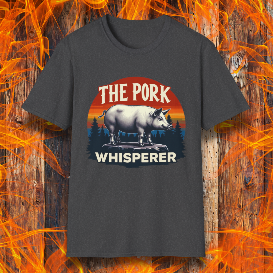 Dark Heather T-shirt featuring a bold and humorous BBQ-themed design. The centerpiece is a detailed pig standing on a wooden stump, framed by a circular emblem with a rustic sunset background in shades of orange, red, and yellow, along with pine tree silhouettes. Above the pig, the text reads "THE PORK WHISPERER" in bold, vintage-style lettering. This design is perfect for BBQ enthusiasts, pitmasters, and pork lovers.