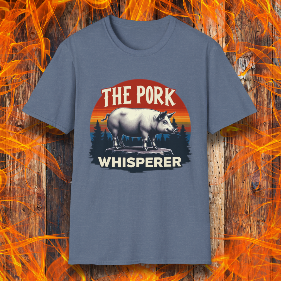 Heather Indigo T-shirt featuring a bold and humorous BBQ-themed design. The centerpiece is a detailed pig standing on a wooden stump, framed by a circular emblem with a rustic sunset background in shades of orange, red, and yellow, along with pine tree silhouettes. Above the pig, the text reads "THE PORK WHISPERER" in bold, vintage-style lettering. This design is perfect for BBQ enthusiasts, pitmasters, and pork lovers.