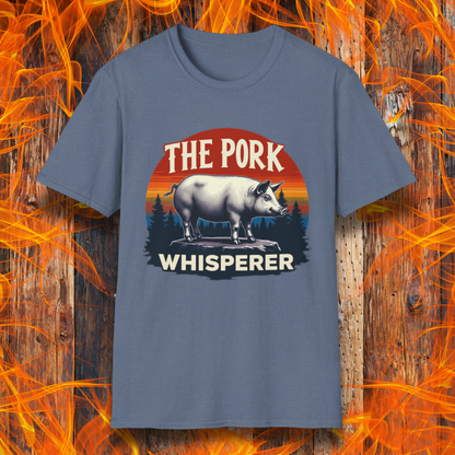 Heather Indigo T-shirt featuring a bold and humorous BBQ-themed design. The centerpiece is a detailed pig standing on a wooden stump, framed by a circular emblem with a rustic sunset background in shades of orange, red, and yellow, along with pine tree silhouettes. Above the pig, the text reads "THE PORK WHISPERER" in bold, vintage-style lettering. This design is perfect for BBQ enthusiasts, pitmasters, and pork lovers.