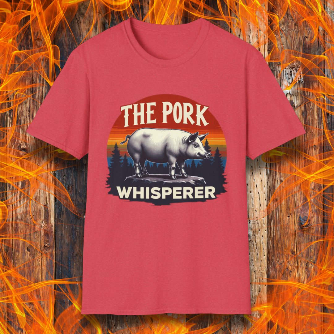 Heather Red T-shirt featuring a bold and humorous BBQ-themed design. The centerpiece is a detailed pig standing on a wooden stump, framed by a circular emblem with a rustic sunset background in shades of orange, red, and yellow, along with pine tree silhouettes. Above the pig, the text reads "THE PORK WHISPERER" in bold, vintage-style lettering. This design is perfect for BBQ enthusiasts, pitmasters, and pork lovers.