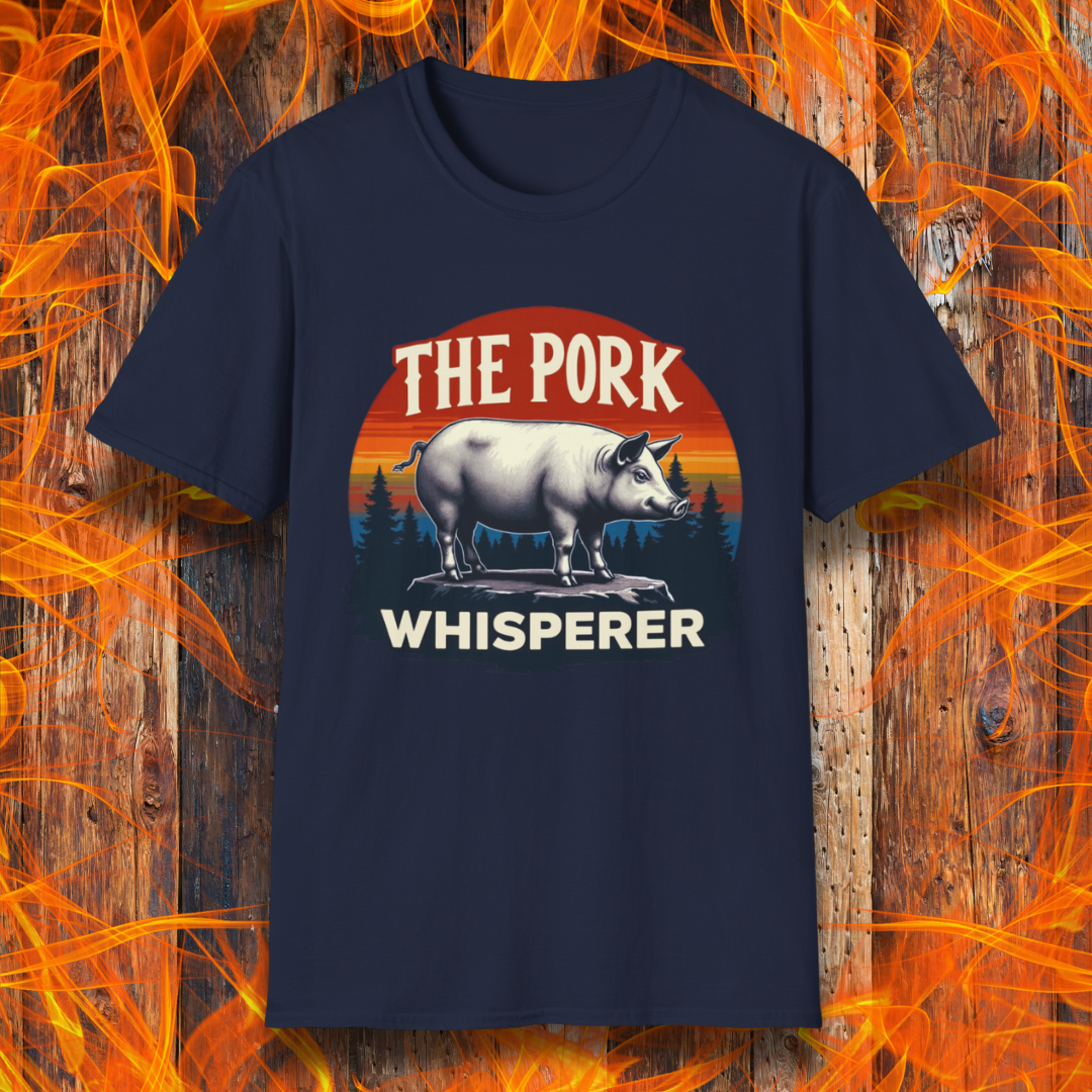 Navy T-shirt featuring a bold and humorous BBQ-themed design. The centerpiece is a detailed pig standing on a wooden stump, framed by a circular emblem with a rustic sunset background in shades of orange, red, and yellow, along with pine tree silhouettes. Above the pig, the text reads "THE PORK WHISPERER" in bold, vintage-style lettering. This design is perfect for BBQ enthusiasts, pitmasters, and pork lovers.
