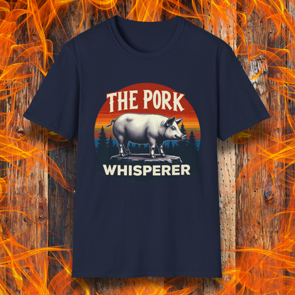 Navy T-shirt featuring a bold and humorous BBQ-themed design. The centerpiece is a detailed pig standing on a wooden stump, framed by a circular emblem with a rustic sunset background in shades of orange, red, and yellow, along with pine tree silhouettes. Above the pig, the text reads "THE PORK WHISPERER" in bold, vintage-style lettering. This design is perfect for BBQ enthusiasts, pitmasters, and pork lovers.