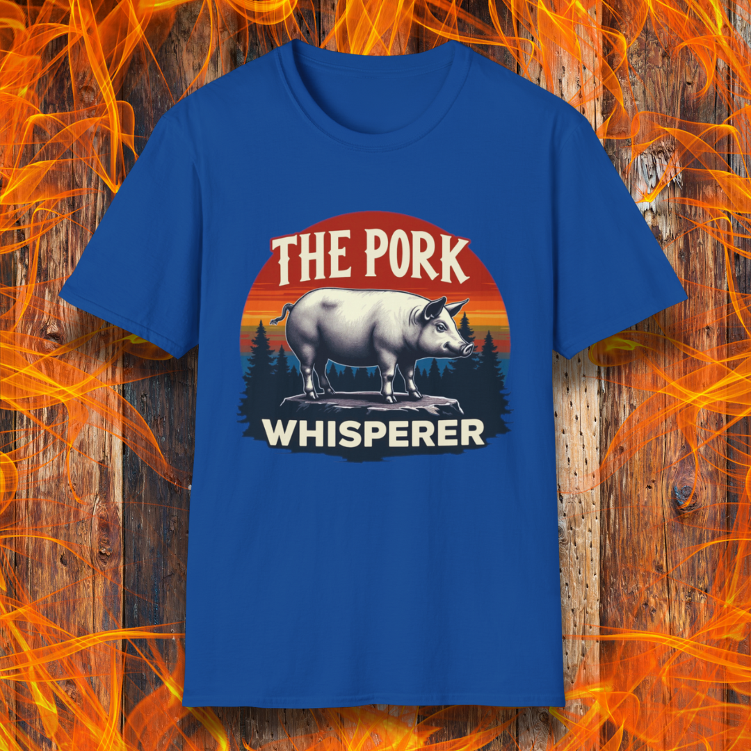 Royal Blue T-shirt featuring a bold and humorous BBQ-themed design. The centerpiece is a detailed pig standing on a wooden stump, framed by a circular emblem with a rustic sunset background in shades of orange, red, and yellow, along with pine tree silhouettes. Above the pig, the text reads "THE PORK WHISPERER" in bold, vintage-style lettering. This design is perfect for BBQ enthusiasts, pitmasters, and pork lovers.