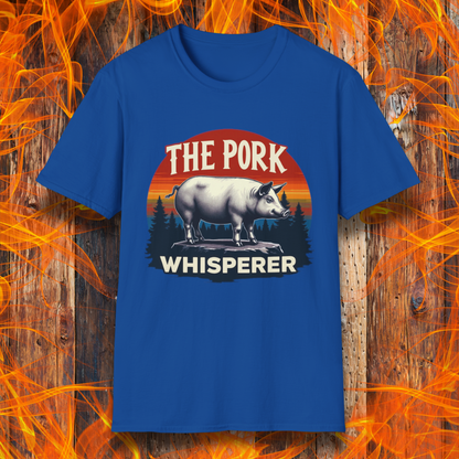 Royal Blue T-shirt featuring a bold and humorous BBQ-themed design. The centerpiece is a detailed pig standing on a wooden stump, framed by a circular emblem with a rustic sunset background in shades of orange, red, and yellow, along with pine tree silhouettes. Above the pig, the text reads "THE PORK WHISPERER" in bold, vintage-style lettering. This design is perfect for BBQ enthusiasts, pitmasters, and pork lovers.