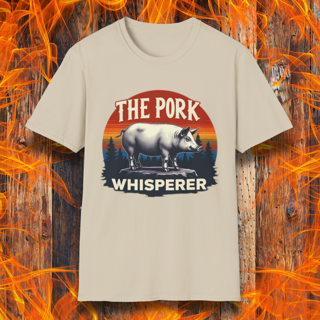 Sand T-shirt featuring a bold and humorous BBQ-themed design. The centerpiece is a detailed pig standing on a wooden stump, framed by a circular emblem with a rustic sunset background in shades of orange, red, and yellow, along with pine tree silhouettes. Above the pig, the text reads "THE PORK WHISPERER" in bold, vintage-style lettering. This design is perfect for BBQ enthusiasts, pitmasters, and pork lovers.