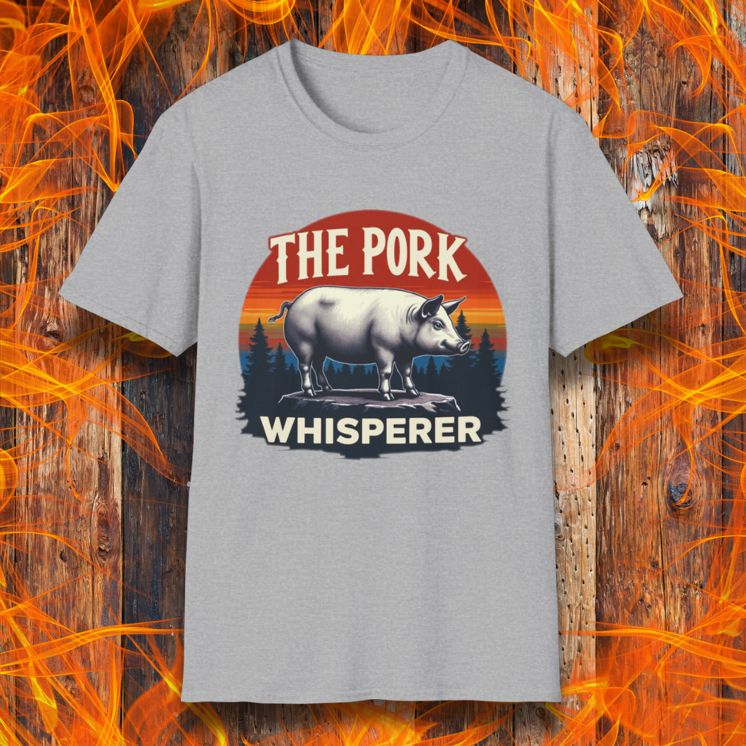 Sport Grey T-shirt featuring a bold and humorous BBQ-themed design. The centerpiece is a detailed pig standing on a wooden stump, framed by a circular emblem with a rustic sunset background in shades of orange, red, and yellow, along with pine tree silhouettes. Above the pig, the text reads "THE PORK WHISPERER" in bold, vintage-style lettering. This design is perfect for BBQ enthusiasts, pitmasters, and pork lovers.