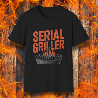 Black T-shirt featuring a bold, red-orange text design that reads "SERIAL GRILLER." Below the text, there's a black and white illustration of a round charcoal grill with flames rising from the grates. The background consists of a rustic wooden surface with bright orange flames surrounding the shirt, creating a fiery grilling-themed aesthetic.