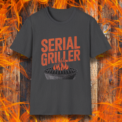 Dark Heather T-shirt featuring a bold, red-orange text design that reads "SERIAL GRILLER." Below the text, there's a black and white illustration of a round charcoal grill with flames rising from the grates. The background consists of a rustic wooden surface with bright orange flames surrounding the shirt, creating a fiery grilling-themed aesthetic.