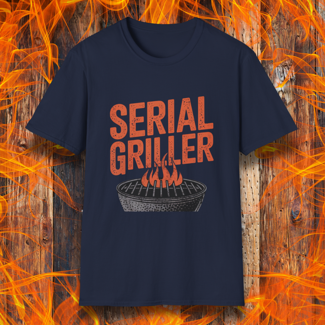 Navy T-shirt featuring a bold, red-orange text design that reads "SERIAL GRILLER." Below the text, there's a black and white illustration of a round charcoal grill with flames rising from the grates. The background consists of a rustic wooden surface with bright orange flames surrounding the shirt, creating a fiery grilling-themed aesthetic.