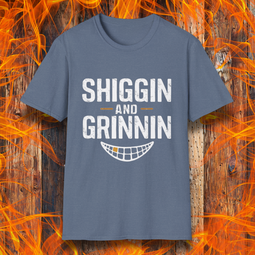 Heather Indigo T-shirt featuring the phrase 'Shiggin and Grinnin' in bold, distressed white lettering. Below the text is a stylized smiling grill grate graphic. The design is set against a rustic wooden background with flames surrounding the edges, giving it a fiery barbecue-themed look.