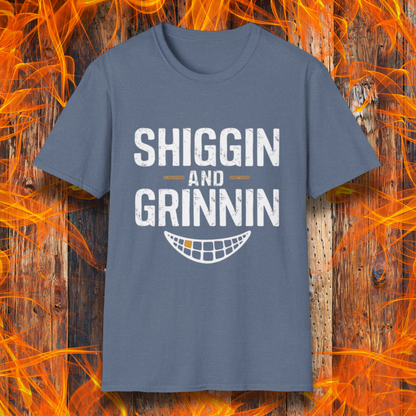 Heather Indigo T-shirt featuring the phrase 'Shiggin and Grinnin' in bold, distressed white lettering. Below the text is a stylized smiling grill grate graphic. The design is set against a rustic wooden background with flames surrounding the edges, giving it a fiery barbecue-themed look.
