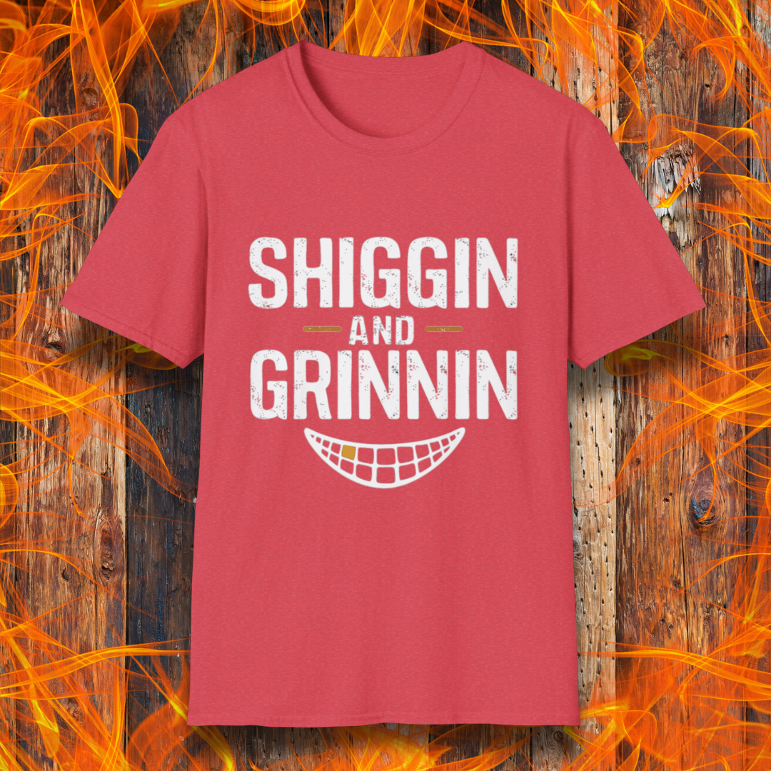 Heather Red T-shirt featuring the phrase 'Shiggin and Grinnin' in bold, distressed white lettering. Below the text is a stylized smiling grill grate graphic. The design is set against a rustic wooden background with flames surrounding the edges, giving it a fiery barbecue-themed look.