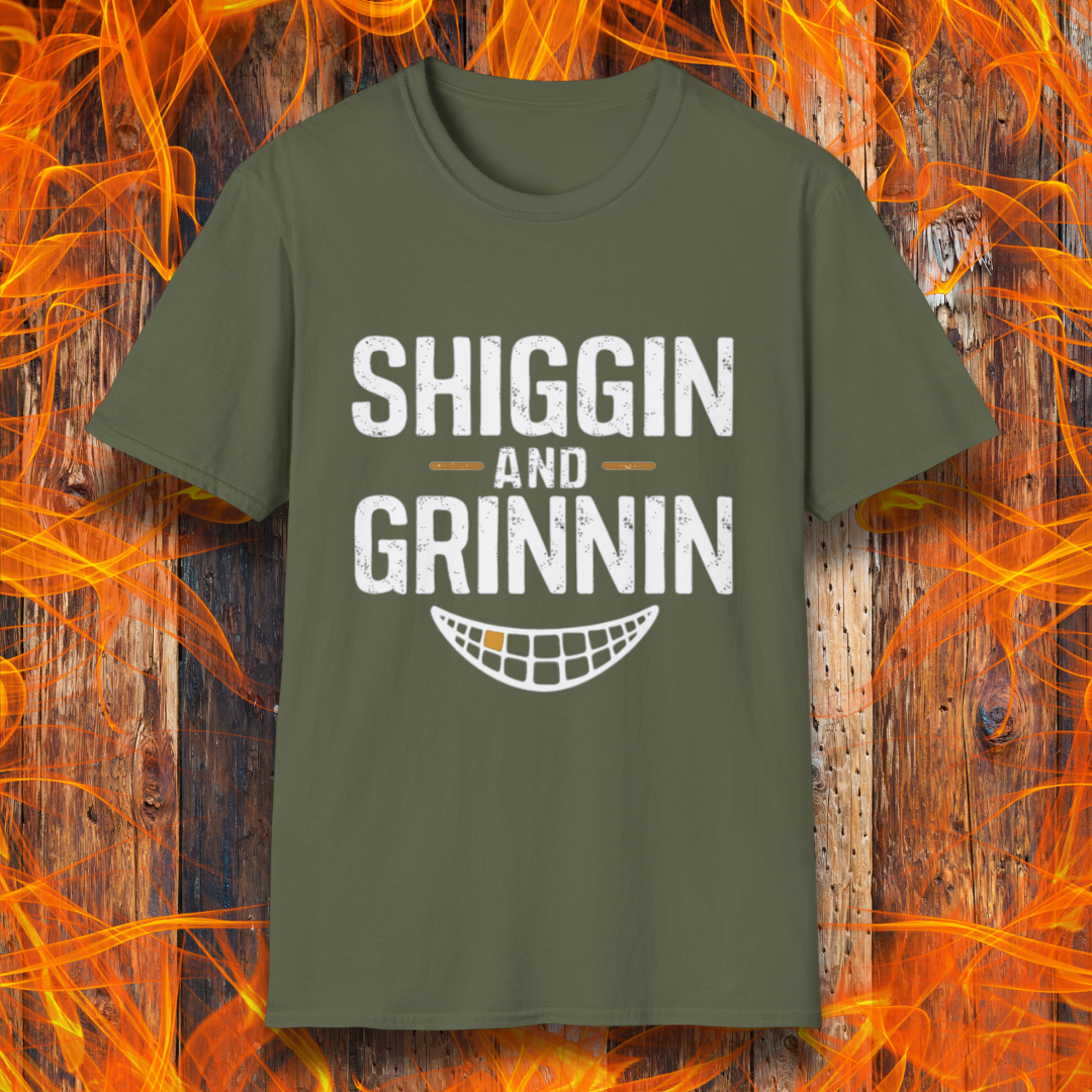 Military Green T-shirt featuring the phrase 'Shiggin and Grinnin' in bold, distressed white lettering. Below the text is a stylized smiling grill grate graphic. The design is set against a rustic wooden background with flames surrounding the edges, giving it a fiery barbecue-themed look.