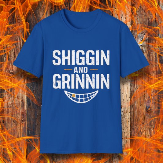 Royal Blue T-shirt featuring the phrase 'Shiggin and Grinnin' in bold, distressed white lettering. Below the text is a stylized smiling grill grate graphic. The design is set against a rustic wooden background with flames surrounding the edges, giving it a fiery barbecue-themed look.