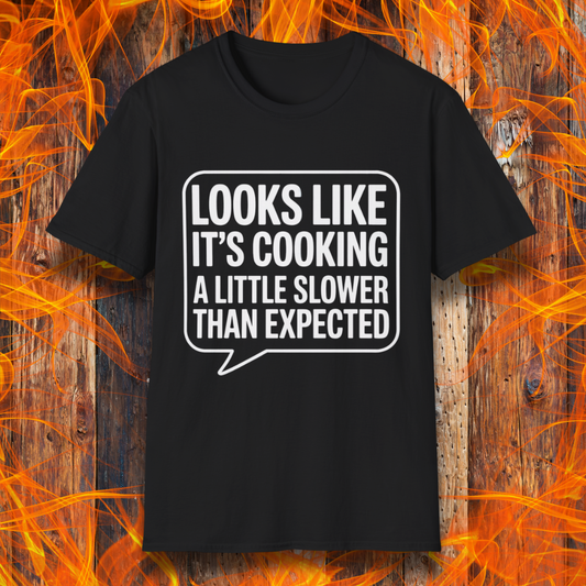 Black T-shirt with white text inside a speech bubble that reads "Looks Like It’s Cooking a Little Slower Than Expected," displayed over a flame and wood background — a humorous BBQ shirt for slow-smoking pros.