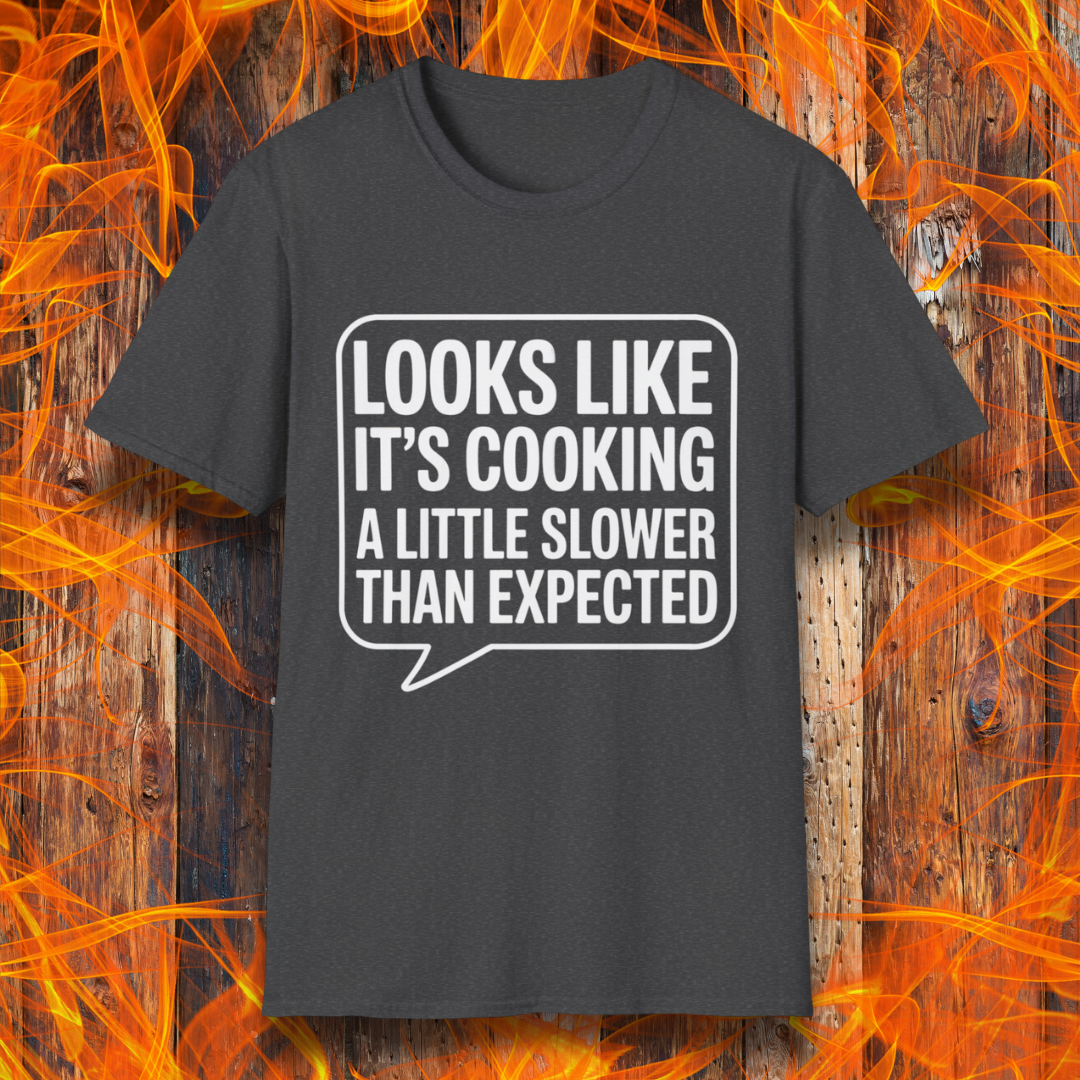 Dark Heather T-shirt with white text inside a speech bubble that reads "Looks Like It’s Cooking a Little Slower Than Expected," displayed over a flame and wood background — a humorous BBQ shirt for slow-smoking pros.