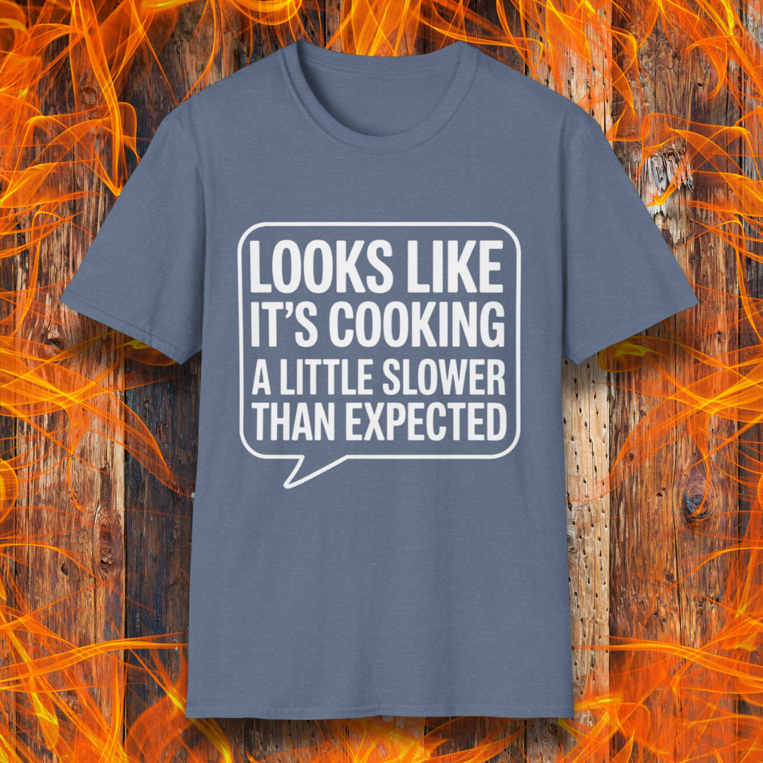 Heather Indigo T-shirt with white text inside a speech bubble that reads "Looks Like It’s Cooking a Little Slower Than Expected," displayed over a flame and wood background — a humorous BBQ shirt for slow-smoking pros.