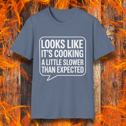 Heather Indigo T-shirt with white text inside a speech bubble that reads "Looks Like It’s Cooking a Little Slower Than Expected," displayed over a flame and wood background — a humorous BBQ shirt for slow-smoking pros.