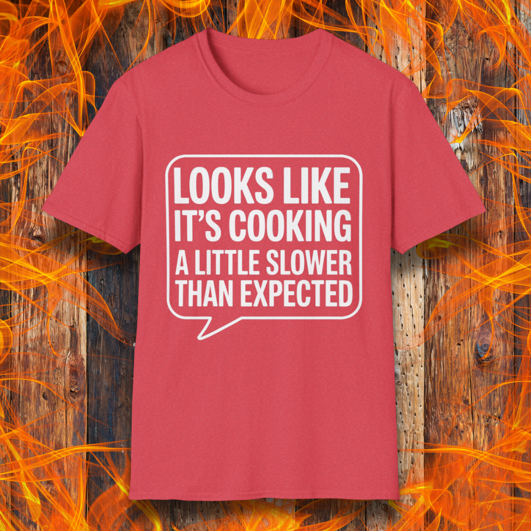 Heather Red T-shirt with white text inside a speech bubble that reads "Looks Like It’s Cooking a Little Slower Than Expected," displayed over a flame and wood background — a humorous BBQ shirt for slow-smoking pros.