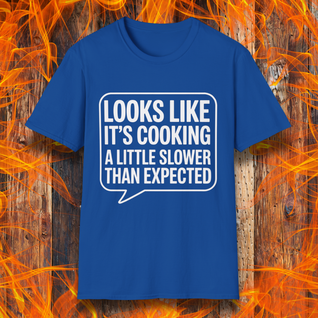 Royal Blue T-shirt with white text inside a speech bubble that reads "Looks Like It’s Cooking a Little Slower Than Expected," displayed over a flame and wood background — a humorous BBQ shirt for slow-smoking pros.