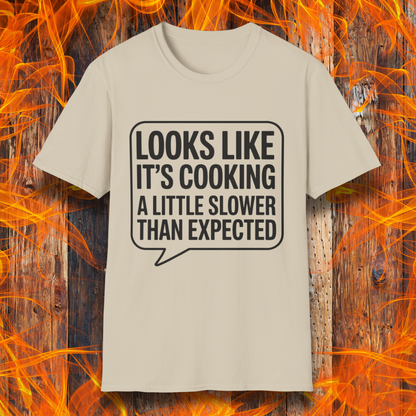 Sand T-shirt with white text inside a speech bubble that reads "Looks Like It’s Cooking a Little Slower Than Expected," displayed over a flame and wood background — a humorous BBQ shirt for slow-smoking pros.