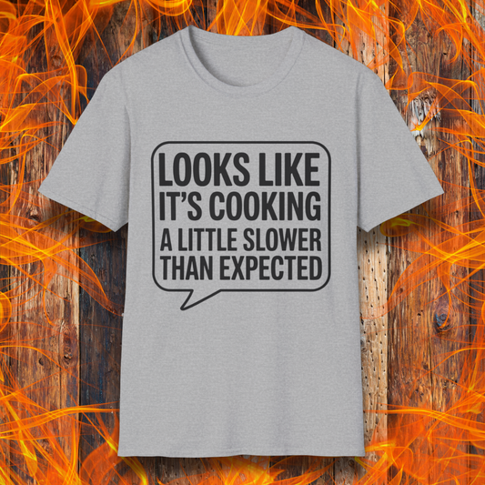 Sport Grey T-shirt with white text inside a speech bubble that reads "Looks Like It’s Cooking a Little Slower Than Expected," displayed over a flame and wood background — a humorous BBQ shirt for slow-smoking pros.